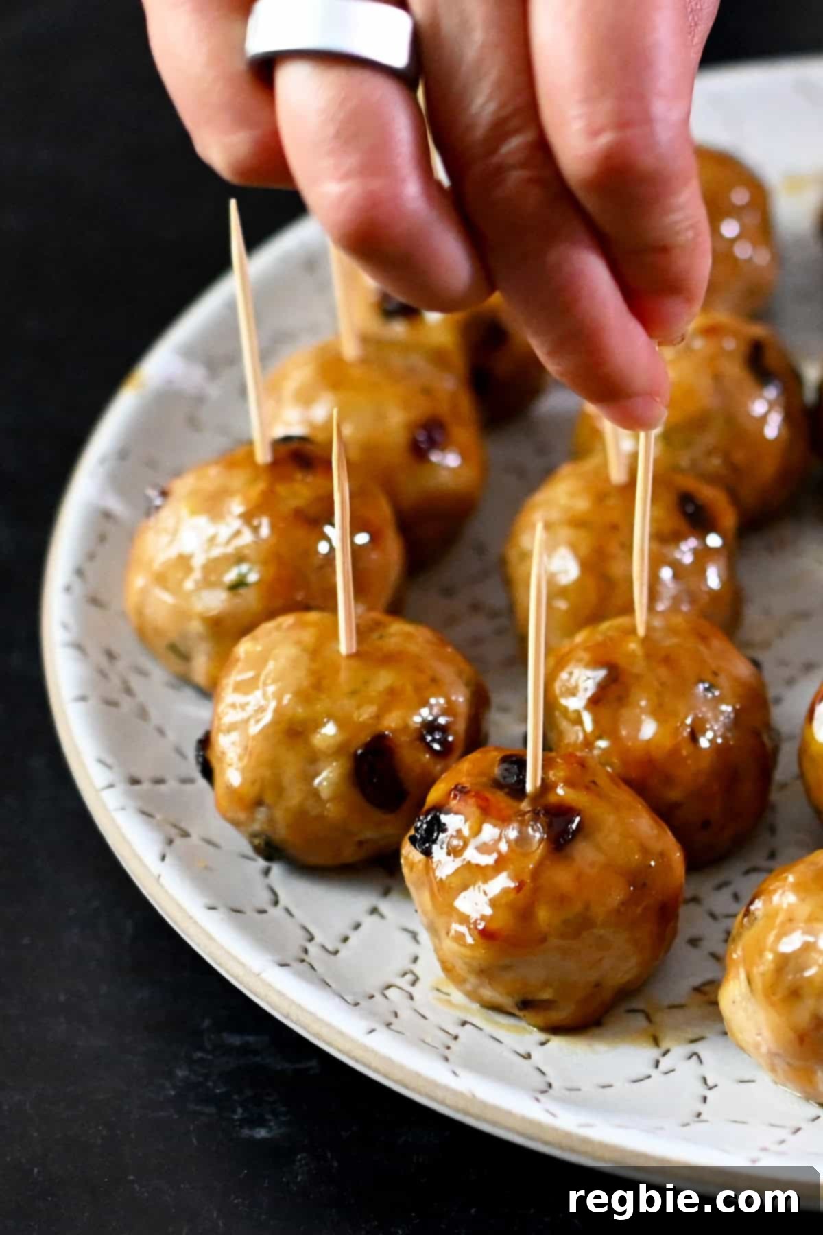 A hand is grabbing a cranberry turkey meatballs topped with a gluten free maple Dijon mustard glaze with a toothpick from a white plate.