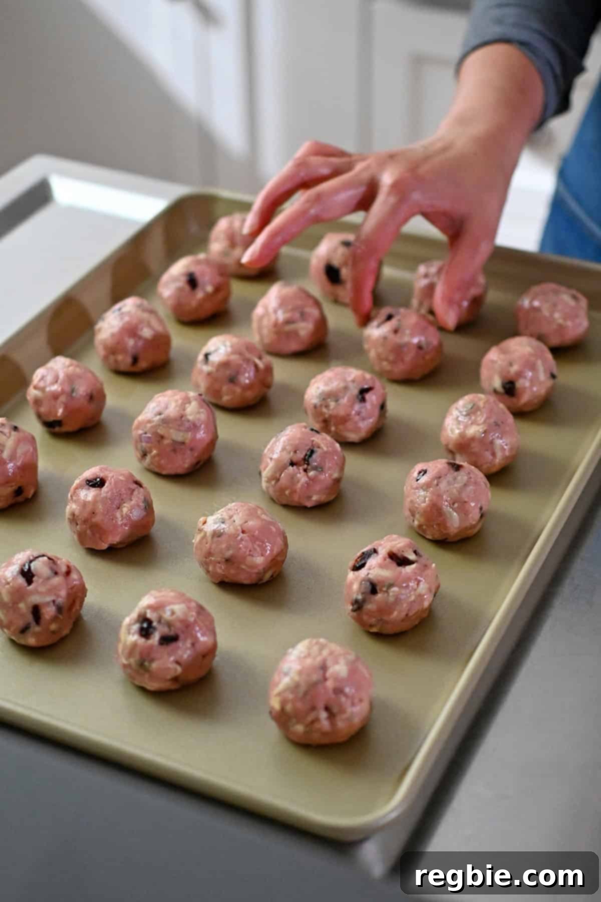A hand is placing raw turkey meatballs with cranberries on a gold rimmed baking sheet.