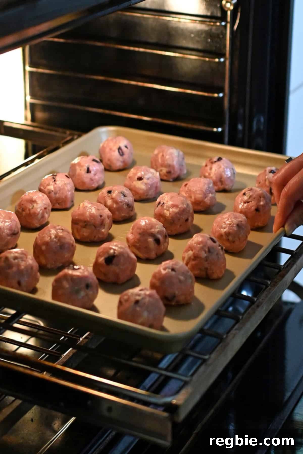 A hand is placing a tray of cranberry turkey meatballs into the oven to cook.