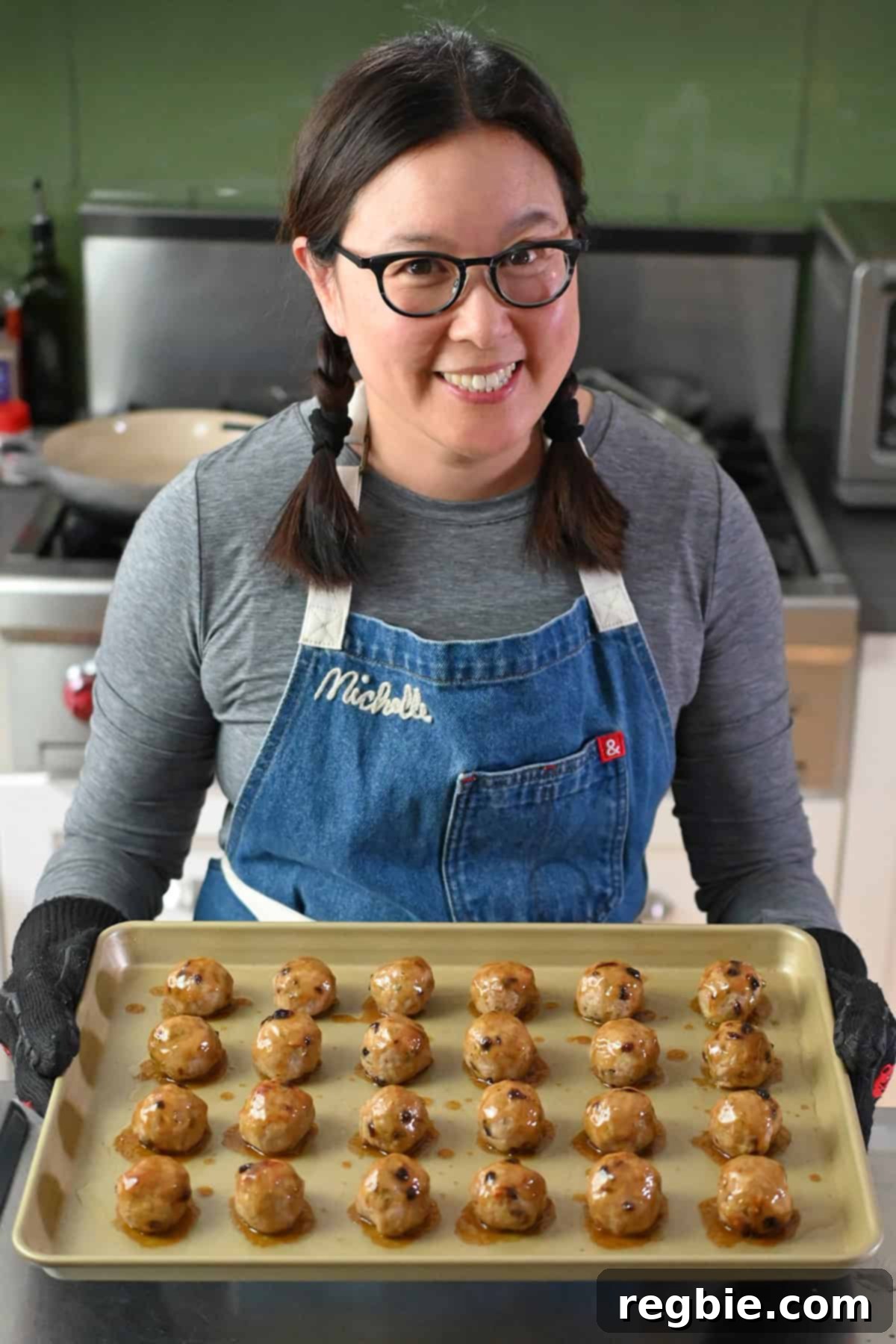A smiling Asian woman in glasses is holding a gold rimmed baking sheet topped with freshly baked cranberry turkey meatballs topped with a maple dijon mustard glaze.