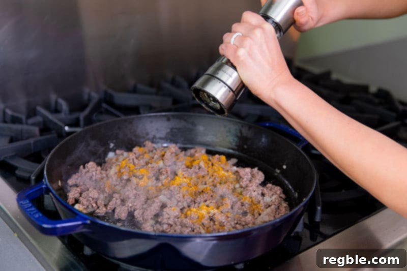 One Pan Beef Cabbage Wonder 11 Someone expertly cracking freshly ground black pepper into a pan filled with savory ground beef, diced onions, and fragrant Madras curry powder, enhancing the flavor.