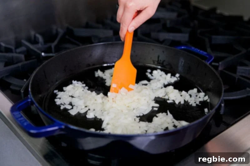 One Pan Beef Cabbage Wonder 7 A vibrant orange silicone spatula gently stirs diced onions in a hot cast iron skillet, cooking them until they become soft and translucent.