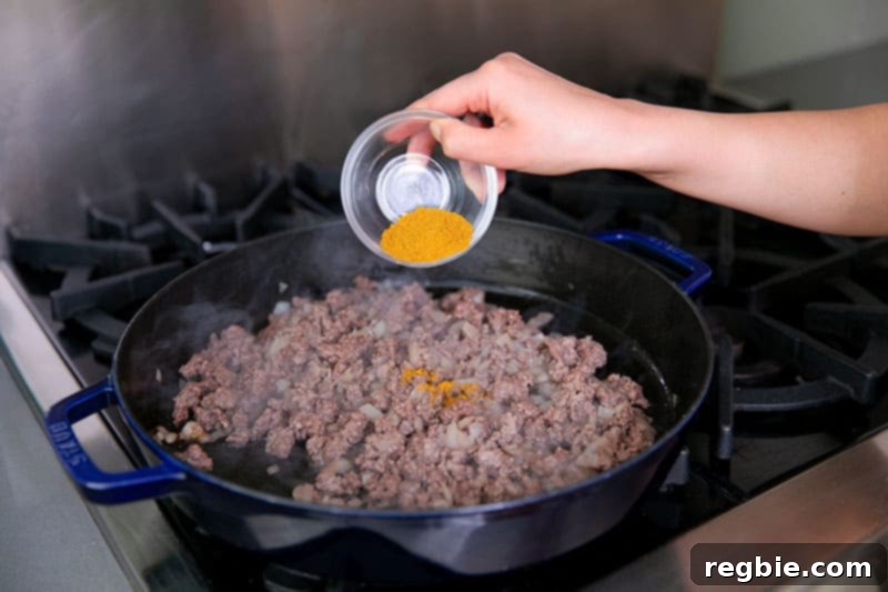 One Pan Beef Cabbage Wonder 10 Pouring a small glass bowl filled with vibrant Indian curry powder directly onto a sizzling cast iron skillet, where ground beef and onions are cooking.