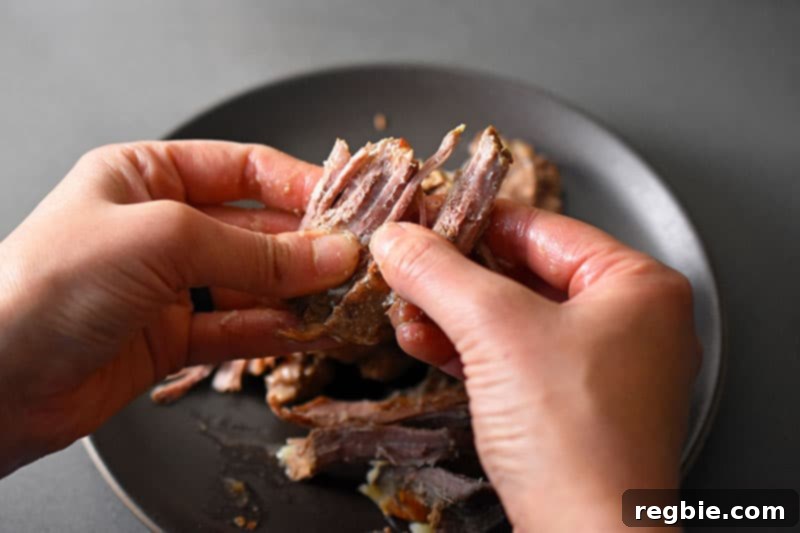 Hands meticulously shredding cold Instant Pot Carnitas by hand into smaller, more manageable pieces, preparing them for the crisping stage.