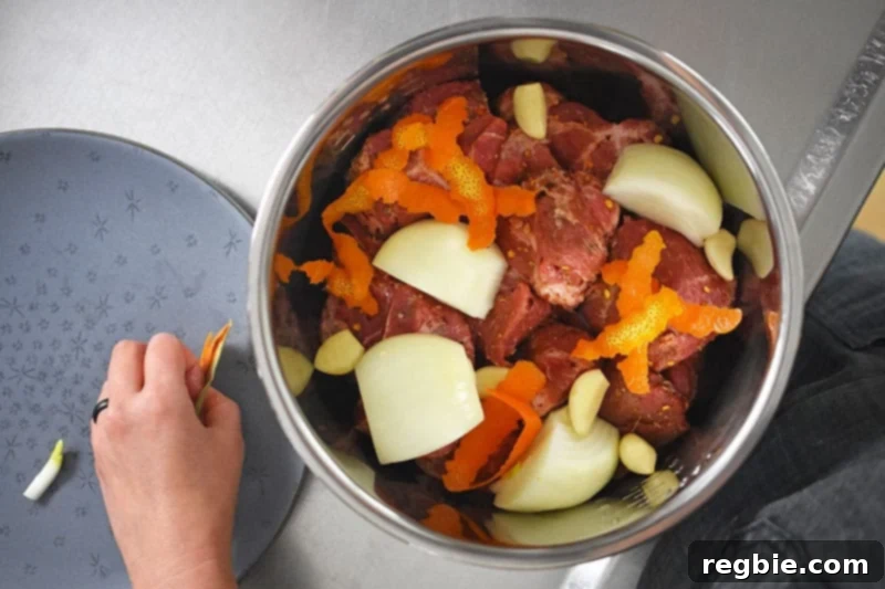 An overhead view inside the Instant Pot insert, showing raw pork cubes nestled with orange zest strips, garlic cloves, quartered yellow onion, and a bay leaf, ready for cooking.