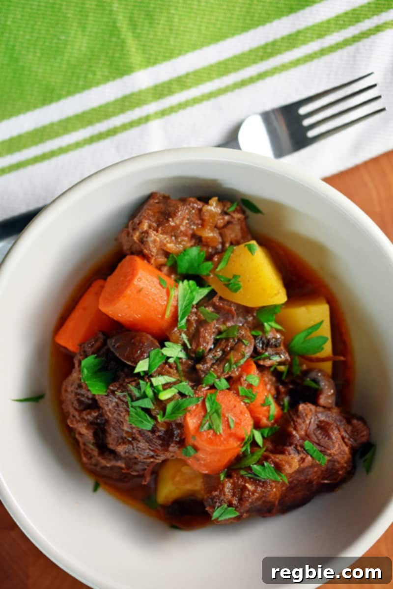 An overhead shot of a white bowl filled with paleo Instant Pot Beef stew with carrots and potatoes. There is a green and white napkin with a fork on top next to the bowl of stew.