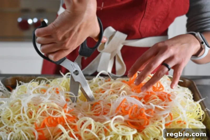 Spiralized white sweet potatoes, carrots, and onions are on a rimmed baking sheet and they are being cut with a pair of kitchen shears.