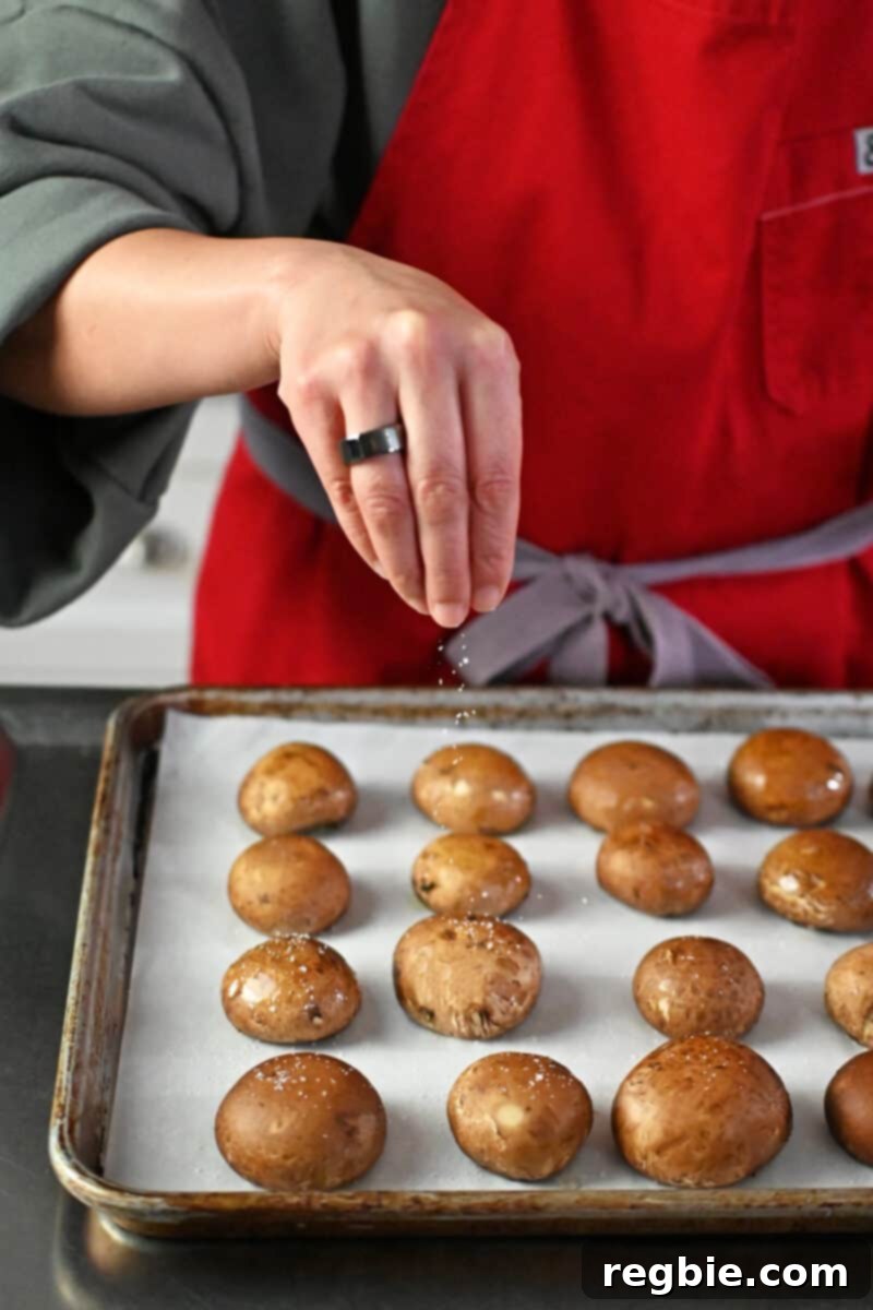Hands carefully sprinkling kosher salt over ghee-coated brown mushrooms, arranged gill-side down on a parchment-lined baking sheet.
