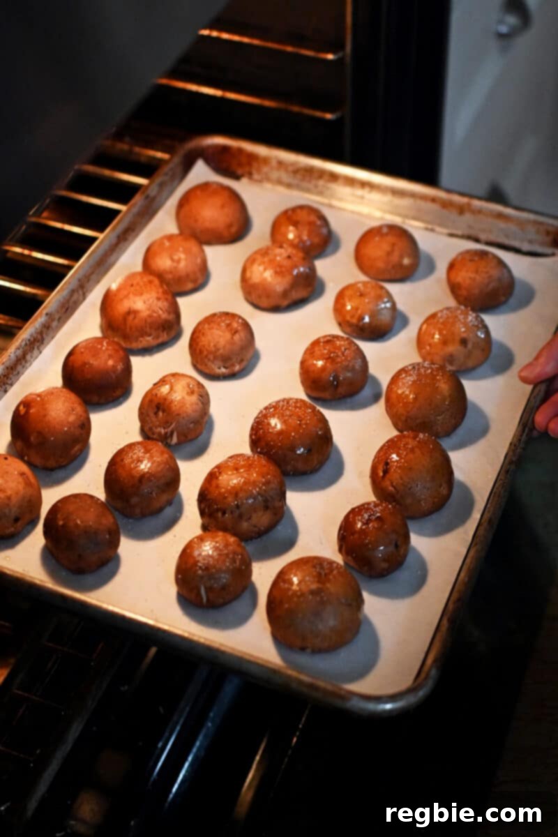 A baking sheet filled with cremini mushrooms, positioned gill-side down, being gently placed into a hot oven for roasting.