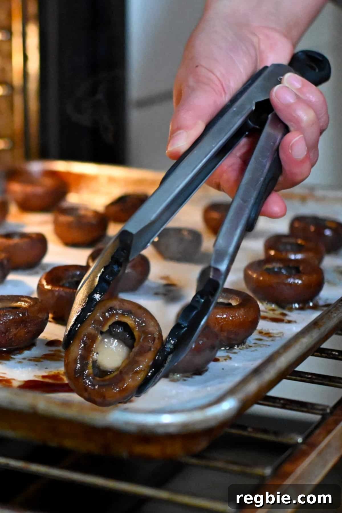A pair of tongs meticulously flipping roasted cremini mushrooms on a baking sheet, turning them gill-side up to continue roasting.