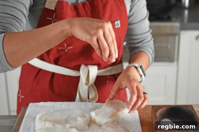 Quick Tomato Poached Cod One-Pan Meal 6 A woman, wearing a vibrant red apron, is depicted carefully sprinkling coarse kosher salt over raw cod fillets. This action highlights the importance of proper seasoning before cooking the fish.