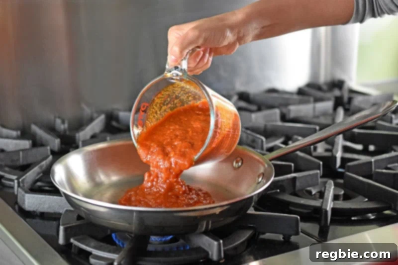 Quick Tomato Poached Cod One-Pan Meal 7 A generous two cups of vibrant marinara sauce is being expertly poured from a clear Pyrex measuring cup into a large, gleaming stainless steel skillet, which rests on a stovetop burner, ready for cooking.