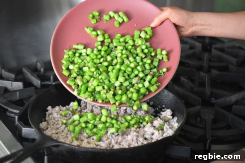 Adding a platter of diced asparagus to the skillet filled with cooked ground pork.