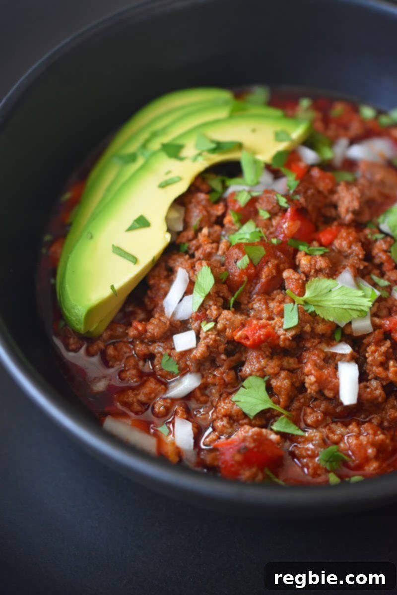 A close-up image of Instant Pot Ground Beef Chili topped with sliced avocado, diced onions, and chopped cilantro in a black bowl, ready to be enjoyed.