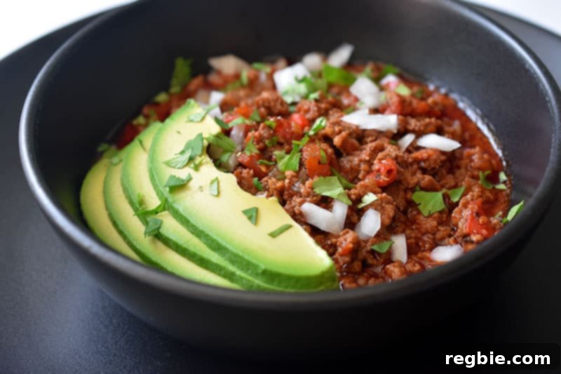 A side view of a black bowl on top of a black plate. The bowl is filled with Instant Pot Ground Beef Chili and topped with sliced avocado, diced white onions, and minced cilantro leaves.