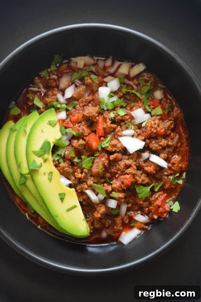 A top view of a black bowl on top of a black plate. The bowl is filled with Instant Pot Ground Beef Chili and topped with sliced avocado, diced white onions, and minced cilantro leaves, presenting a vibrant and appetizing meal.