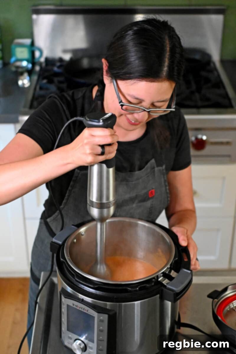 A smiling Asian woman in glasses is using an immersion blender to blend a sauce in an Instant Pot.