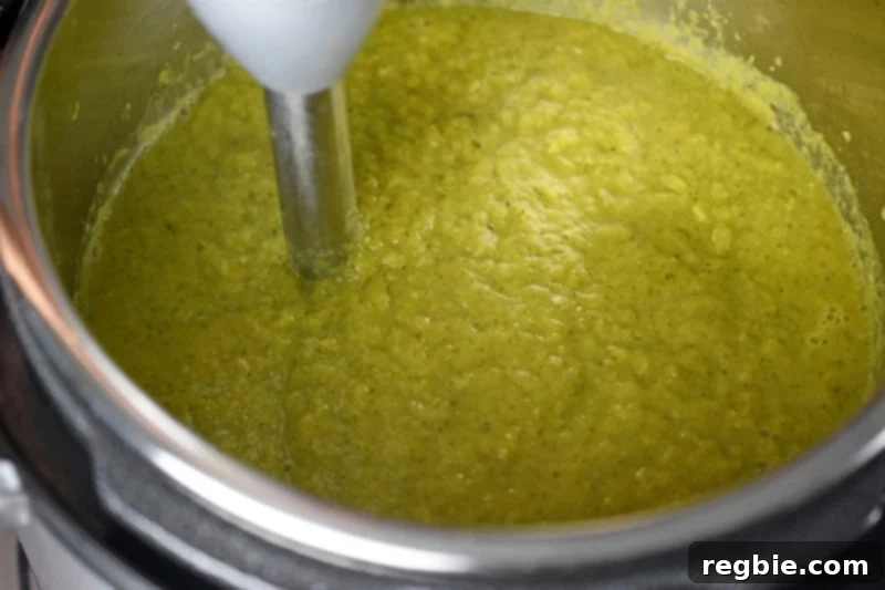 An immersion blender actively pureeing the cooked vegetables directly inside the Instant Pot, transforming them into a smooth, vibrant green Curried Cream of Broccoli Soup.