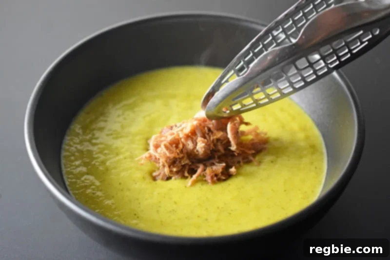 A bowl of rich Instant Pot Curried Cream of Broccoli Soup being topped generously with shredded, crisped Kalua Pig, adding a hearty and flavorful element to the meal.