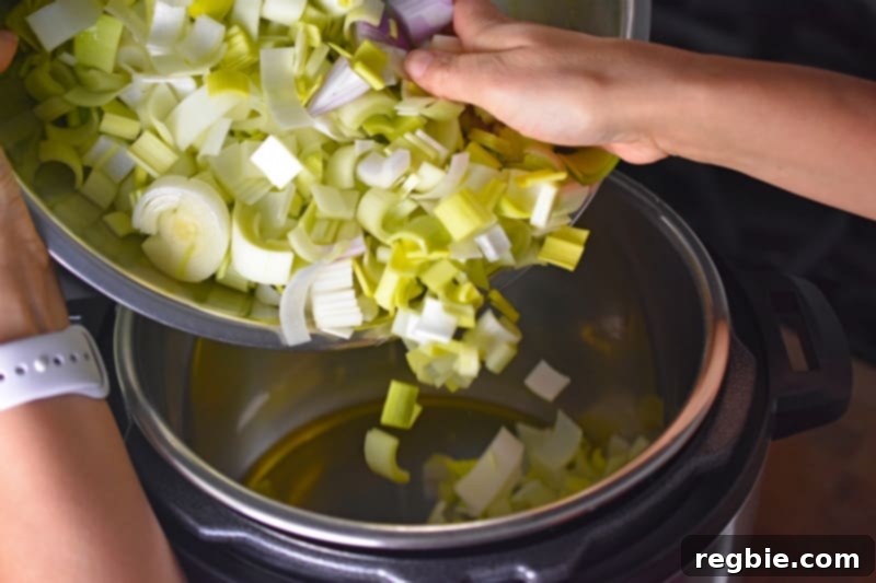 A hand adding chopped leeks and shallots to a hot Instant Pot with cooking fat, beginning the flavorful base for Whole30 Curried Cream of Broccoli Soup.