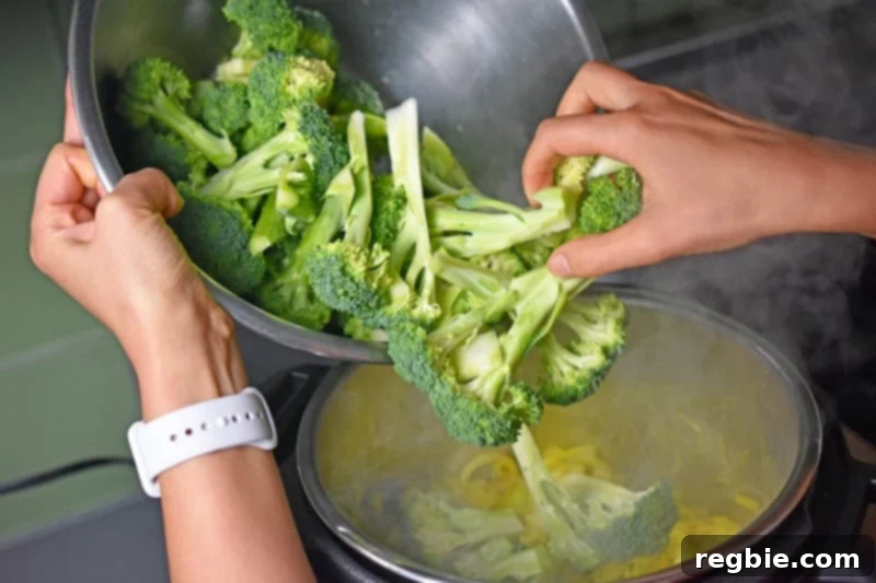 A generous amount of chopped broccoli florets being added to the Instant Pot, joining the sautéed aromatics for the Curried Cream of Broccoli Soup.