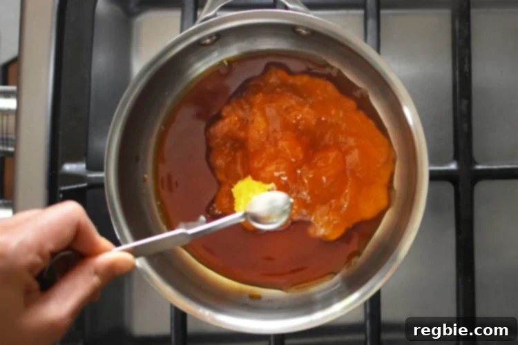 An overhead shot of someone adding a spoonful of minced ginger to a saucepan filled with ingredients for homemade Whole30 Apricot-Ginger Sauce.