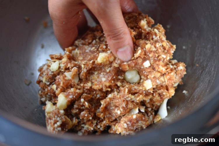 A close-up view of hands expertly mixing the sticky date, coconut, and macadamia nut mixture within a metal bowl, diligently working to form the homogeneous base for the homemade Liar Balls.