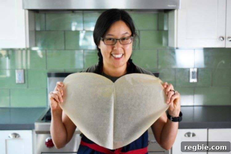 A hand holding up a heart-shaped piece of parchment paper, demonstrating the preparation for Fiona’s Phenomenal Fish Packets.