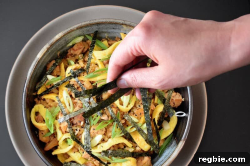 A bowl of paleo kimchi cauliflower fried rice being garnished with thin strips of toasted seaweed (nori) for a final flourish.