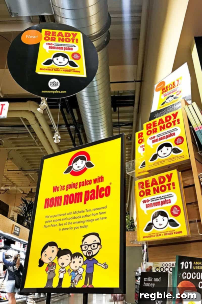 Michelle Tam of Nom Nom Paleo in a Whole Foods Market aisle, pointing at a Nom Nom Paleo Picks sign, wearing a distinctive apron and smile.