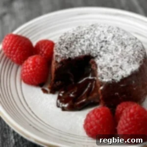 A tantalizing close-up shot of a white plate featuring a chocolate lava cake, artfully cut open to reveal its irresistible gooey, molten center. The cake is dusted with powdered keto confectioners' sugar and garnished with fresh raspberries, promising a truly decadent experience.