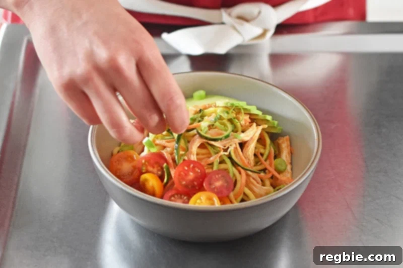 A beautifully presented bowl of spicy Thai chicken zoodle salad, richly garnished with fresh tomatoes, creamy avocado, and aromatic herbs.