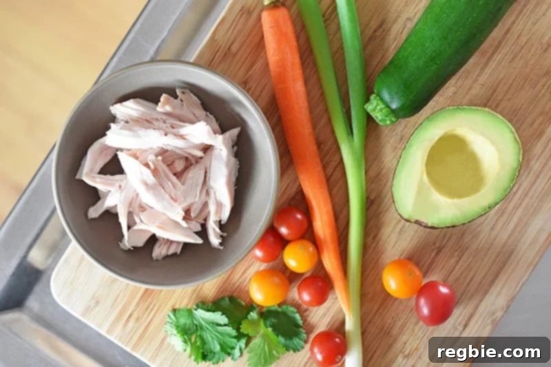 An array of fresh ingredients for the spicy Thai zoodle salad laid out on a countertop, including zucchini, carrots, and shredded chicken, ready for preparation.