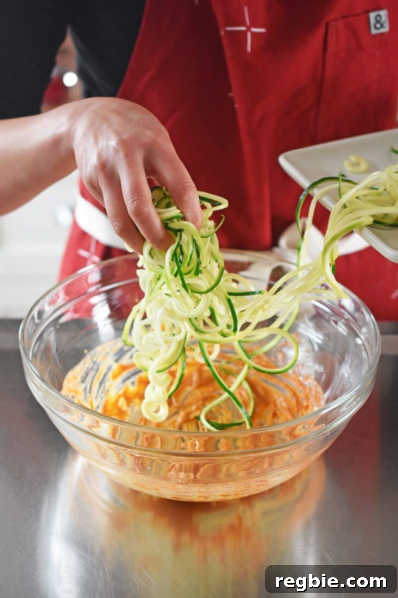 Raw spiralized zucchini noodles being gently added to a large bowl already containing the spicy Thai dressing.