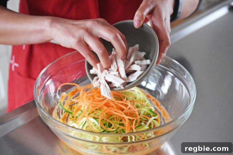 Shredded cooked chicken being added to a bowl with julienned vegetables and dressing, ready for mixing to create the spicy Thai zoodle salad.