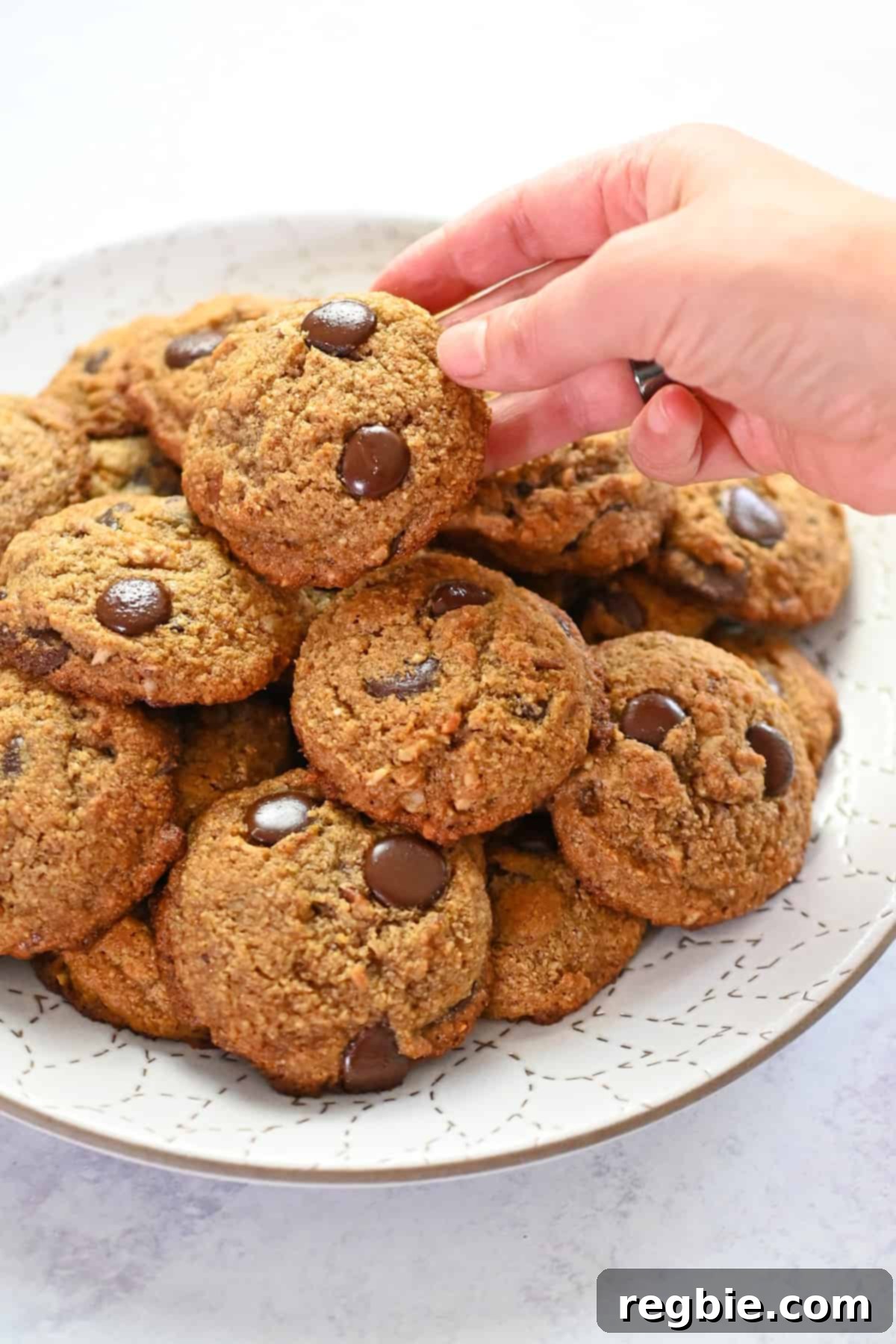 A hand is gently grabbing a soft paleo banana chocolate chip cookie from a white plate piled high with more cookies.