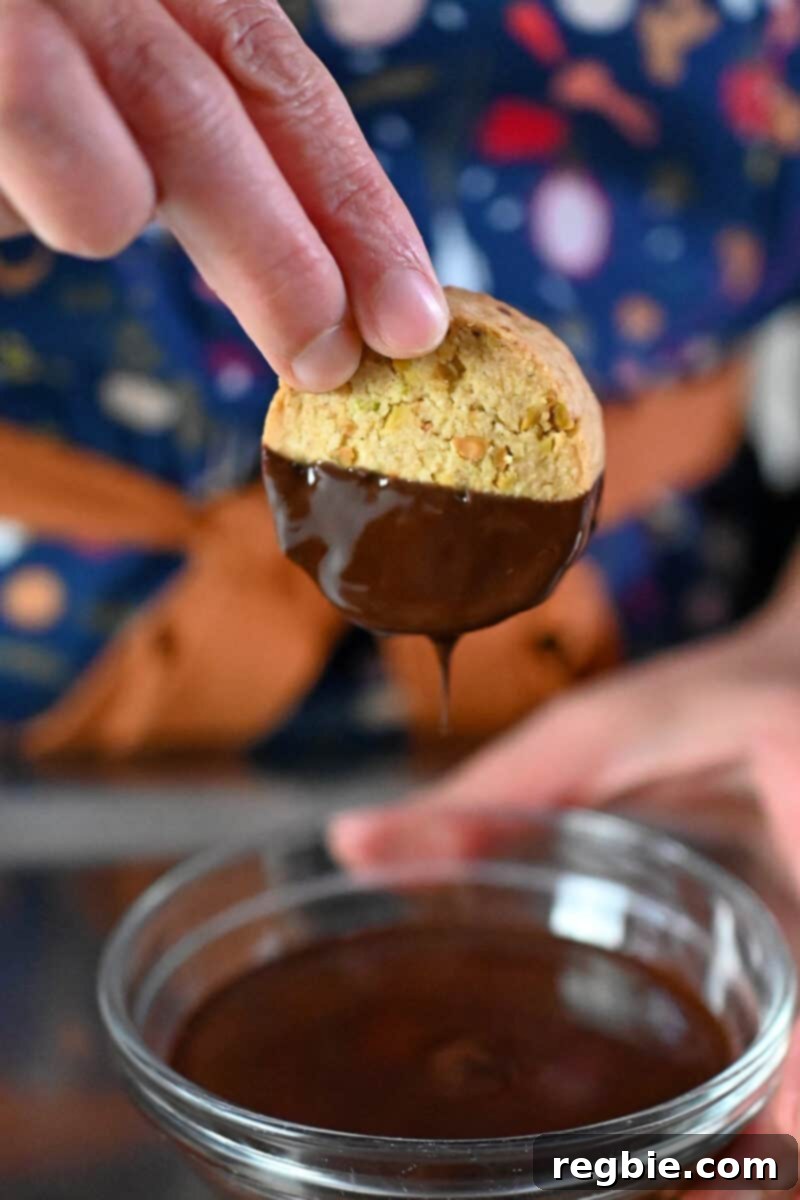 A hand is dunking a crunchy pistachio cookie into a bowl filled with melted dark chocolate, ready for gifting.
