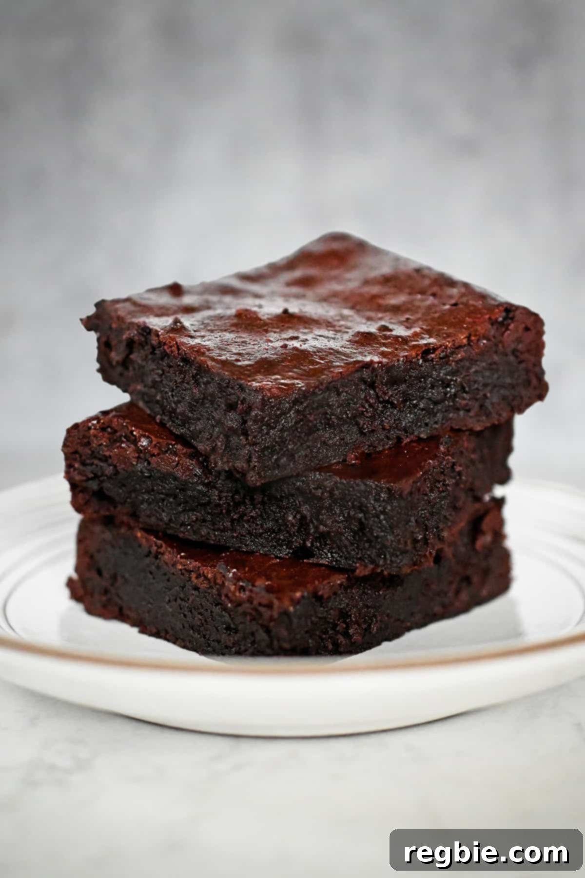 A pristine white plate is stacked with three perfectly cut squares of fudgy paleo brownies.