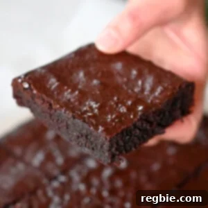A hand is holding a square of fudgy paleo brownies above a cutting board filled with the remaining delicious paleo brownies.