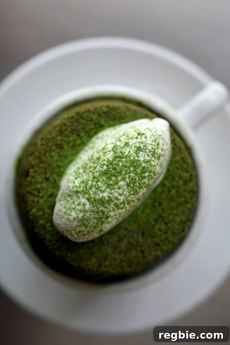 An overhead shot of a white coffee mug filled with a vibrant gluten-free matcha mug cake, topped with fluffy coconut whipped cream and dusted with matcha powder.