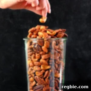 A hand reaching for a cashew from a tall glass jar filled with a delicious assortment of paleo spiced nuts.