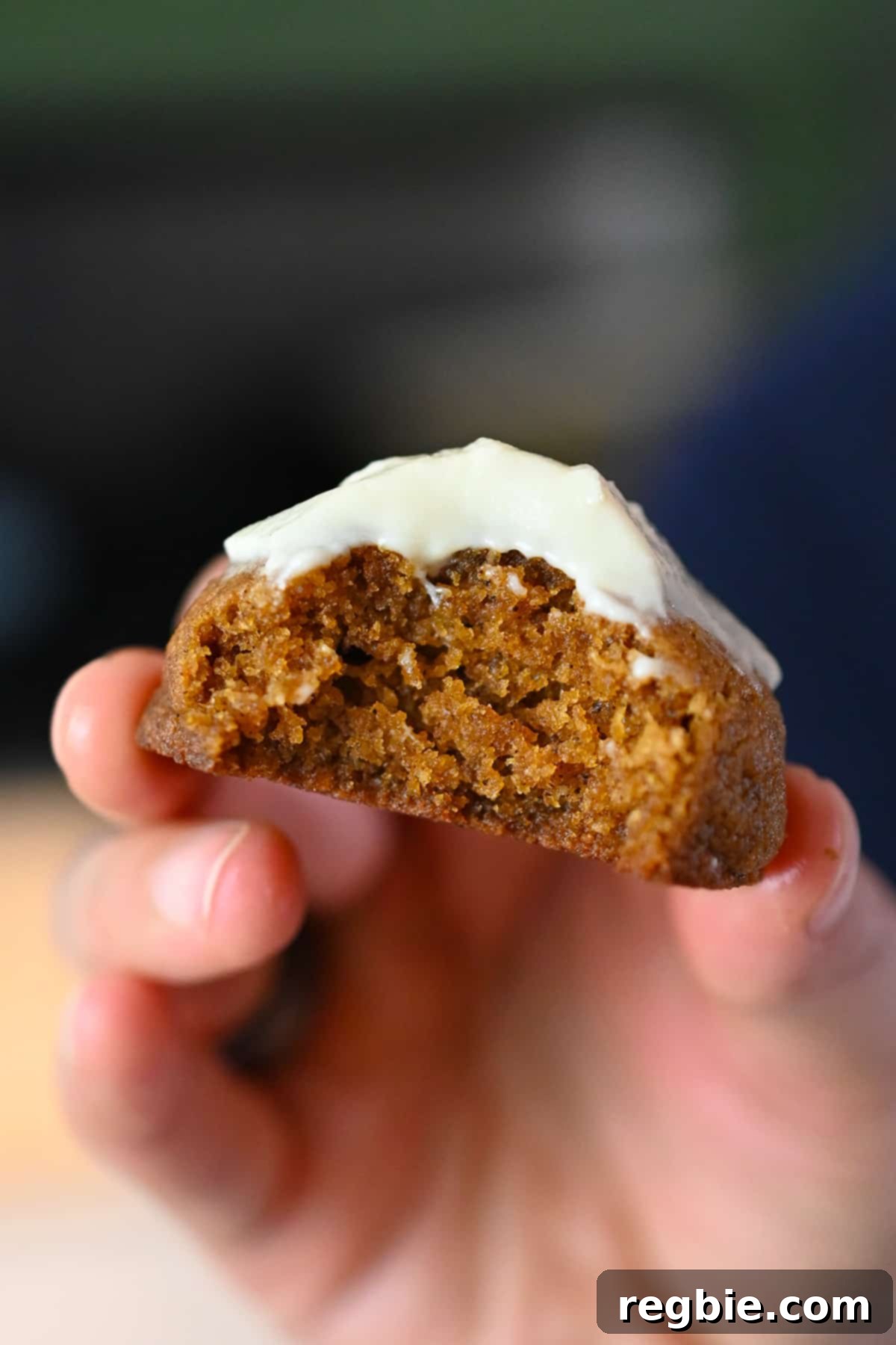A hand holding a frosted paleo pumpkin cookie with a bite taken, revealing its moist, cake-like texture.