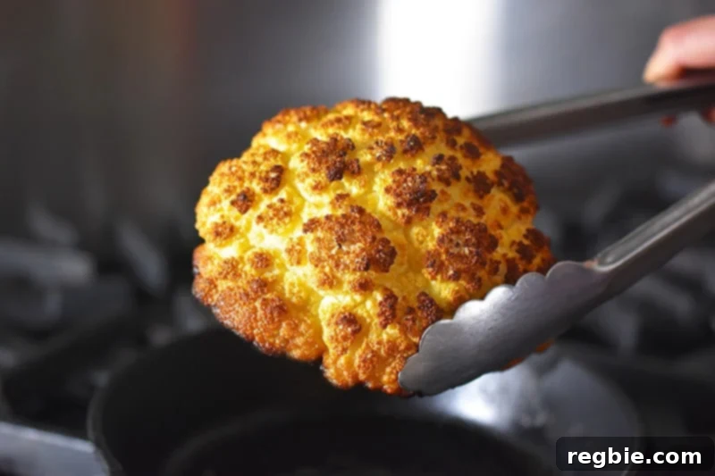 A pair of tongs carefully removing the Whole Roasted Cauliflower from the cast iron skillet onto a serving platter.