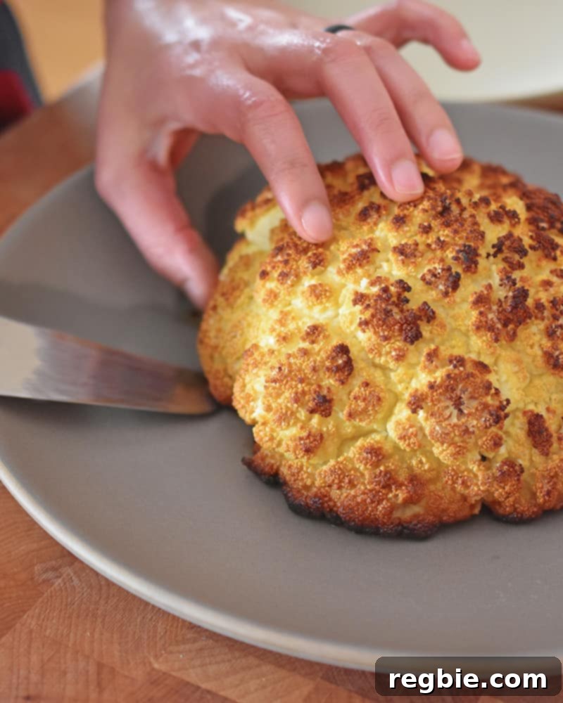 A close-up shot of someone slicing a perfectly cooked Whole Roasted Cauliflower on a gray plate.