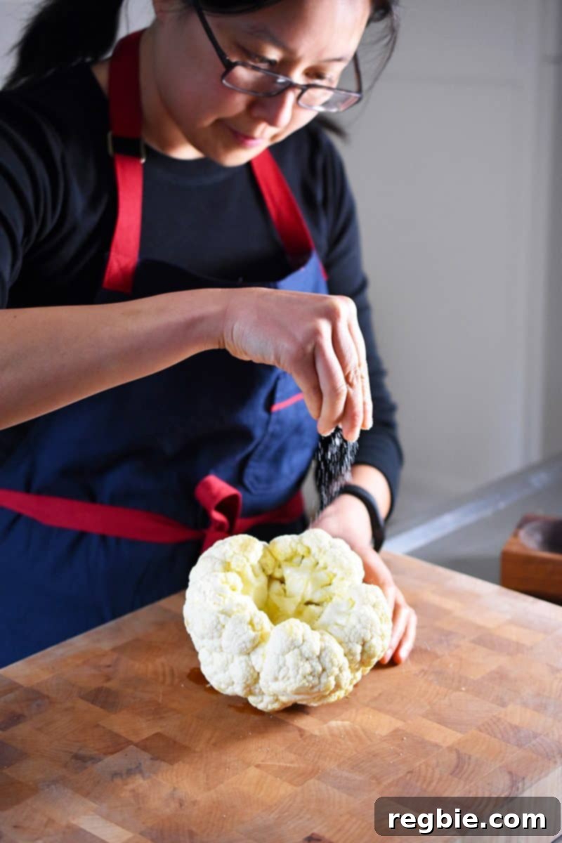 A woman's hands are sprinkling a generous amount of salt all over a whole cauliflower head coated with olive oil.