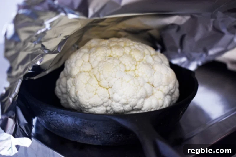 The seasoned cauliflower head is resting in a small cast iron skillet, tightly covered with aluminum foil.