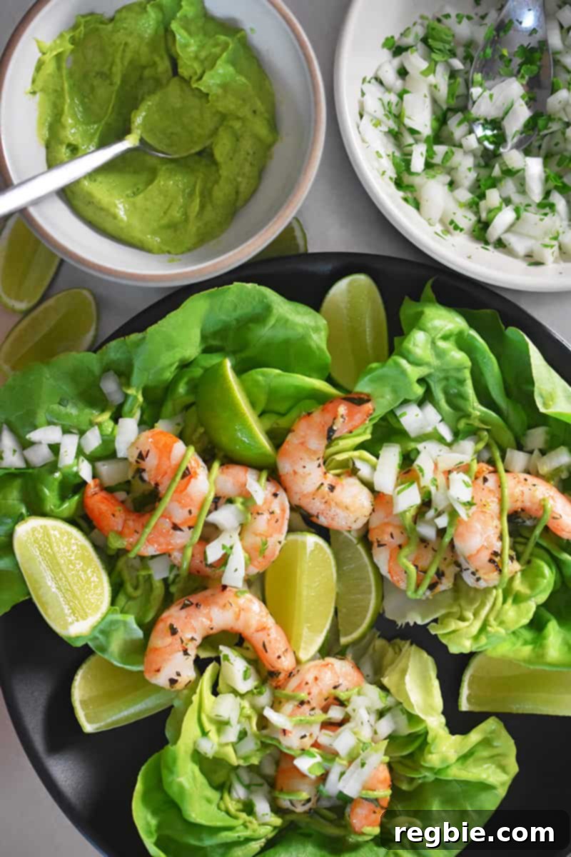 Paleo shrimp tacos arranged on a bed of fresh lettuce, served on a plate with vibrant lime slices. Surrounding the plate are bowls of creamy guacamole and finely chopped onions, adding to the fresh appeal.