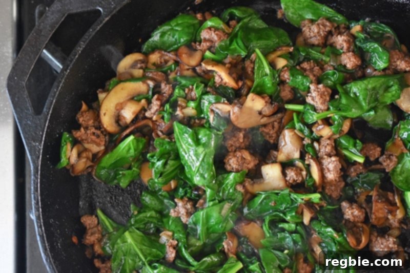 An overhead view of the skillet showcasing cooked ground beef, mushrooms, and vibrant spinach, ready for the eggs.