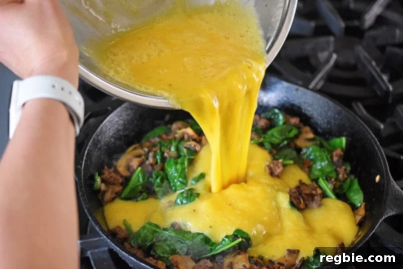 A bowl of whisked scrambled eggs is poured into the pan, mixing with the cooked ground beef, spinach, mushrooms, and onion.