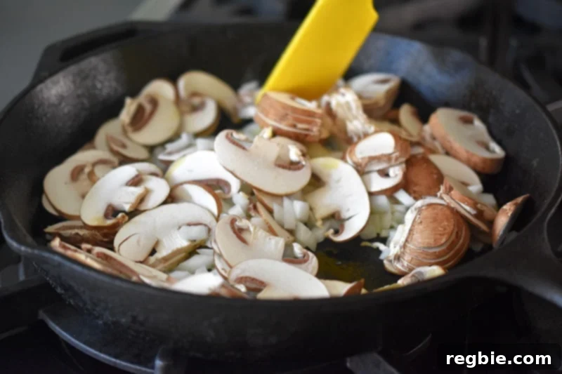 A yellow spatula stirs mushrooms and onions in a large cast iron skillet, ensuring even browning and rich flavor development.